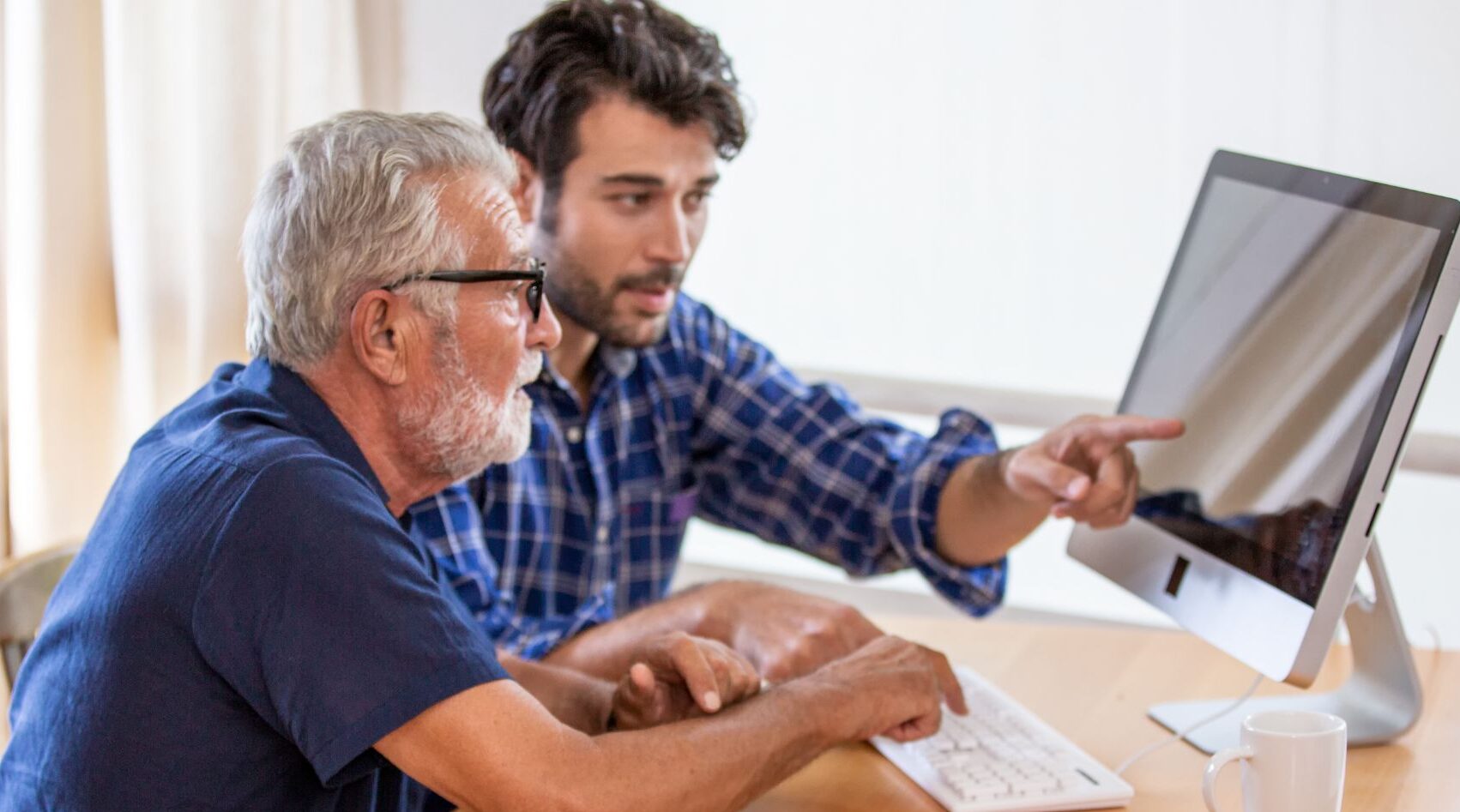 Tecnologia para idosos: homem idoso aprendendo a utilizar um computador, com um homem mais jovem