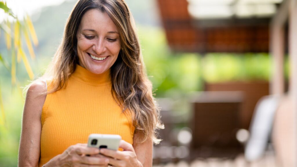 Mulher na rua com celular na mão, olhando pra ele e sorrindo. Ela busca apps para melhorar sua saúde.